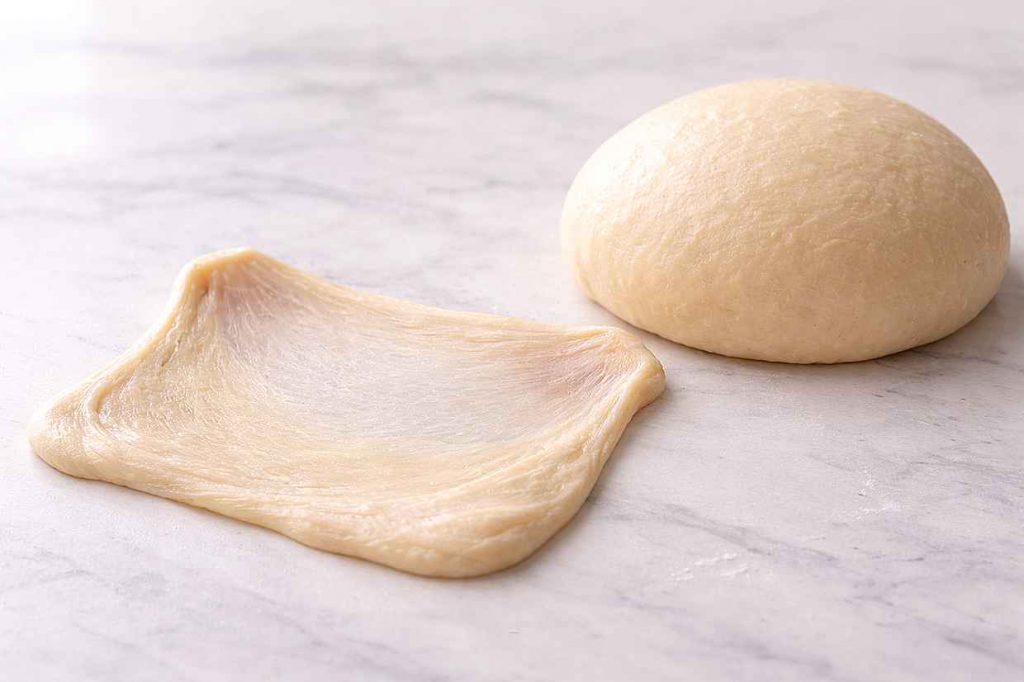 Small piece of smooth bread dough stretched into a thin translucent membrane on a white marble surface showing fully developed gluten