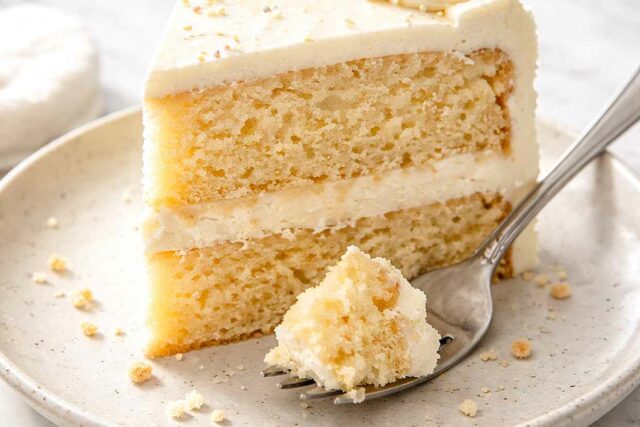 Slice of vanilla layer cake with white buttercream frosting on a speckled ceramic plate with a fork and crumbs showing the soft golden crumb layers