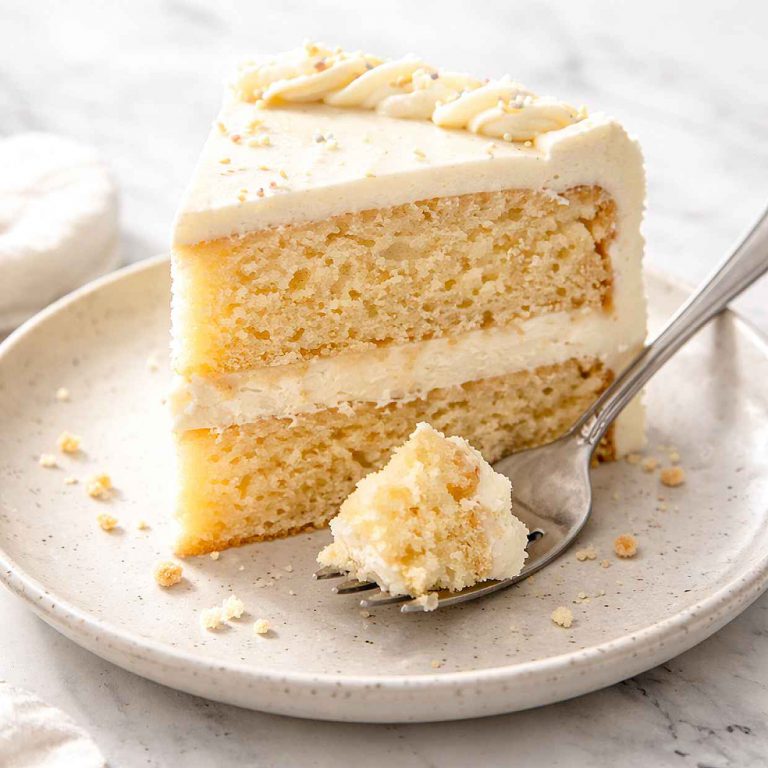 Slice of vanilla layer cake with white buttercream frosting on a speckled ceramic plate with a fork and crumbs showing the soft golden crumb layers