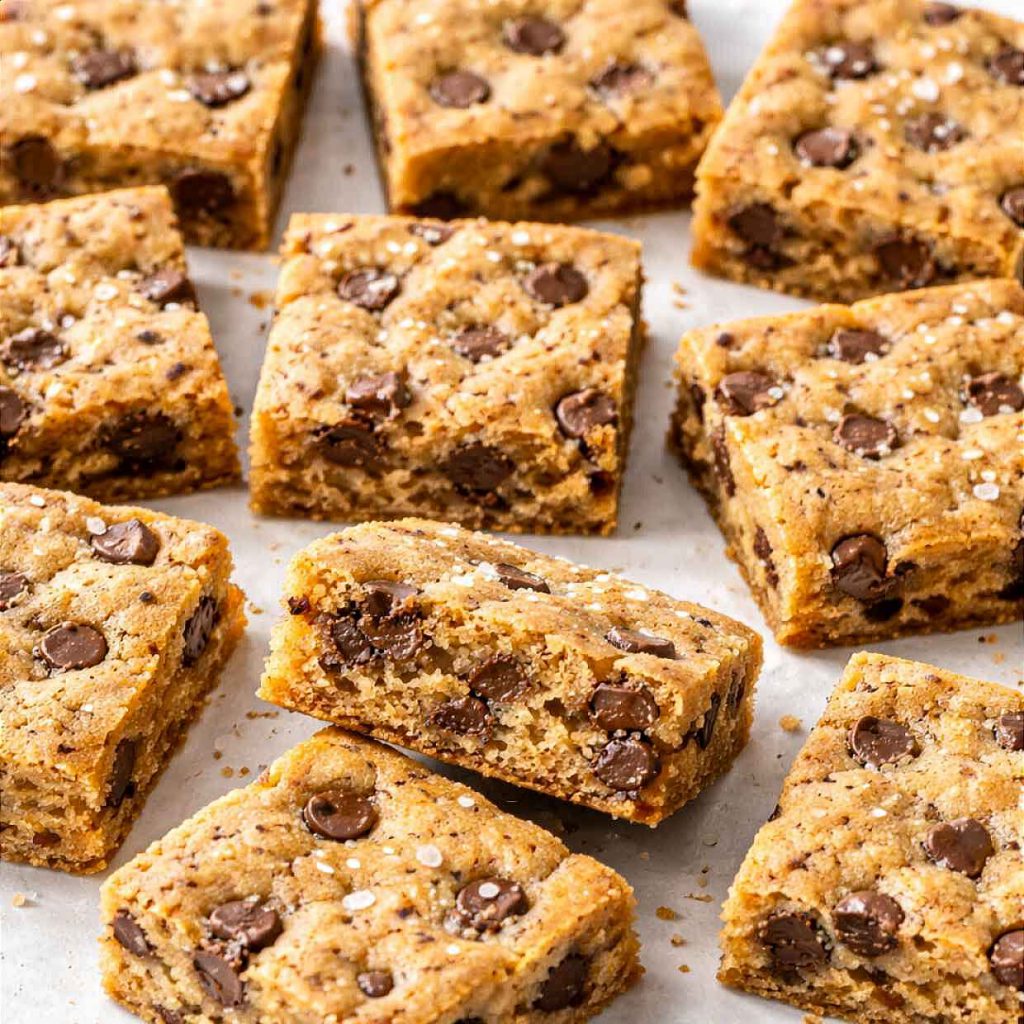 Chocolate chip cookie bars cut into squares scattered on white parchment paper showing golden chewy centers with chocolate chips and sea salt flakes on top