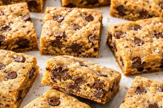 Chocolate chip cookie bars cut into squares scattered on white parchment paper showing golden chewy centers with chocolate chips and sea salt flakes on top