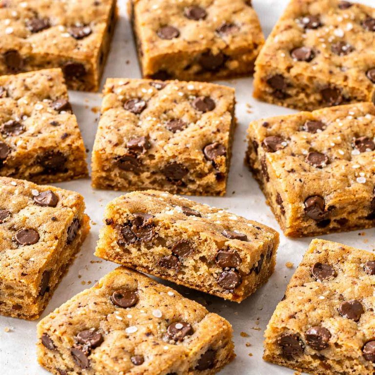 Chocolate chip cookie bars cut into squares scattered on white parchment paper showing golden chewy centers with chocolate chips and sea salt flakes on top