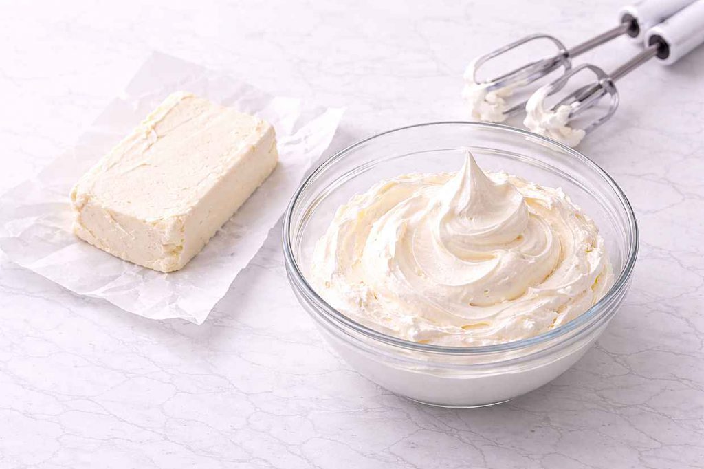 Block of cream cheese at room temperature beside a glass bowl of whipped cream on a white ceramic surface showing correct no-bake ingredient preparation