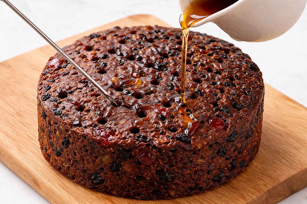 Whole dark Christmas fruitcake on a wooden board being fed with brandy poured from a small ceramic jug over skewer holes in the top of the cake