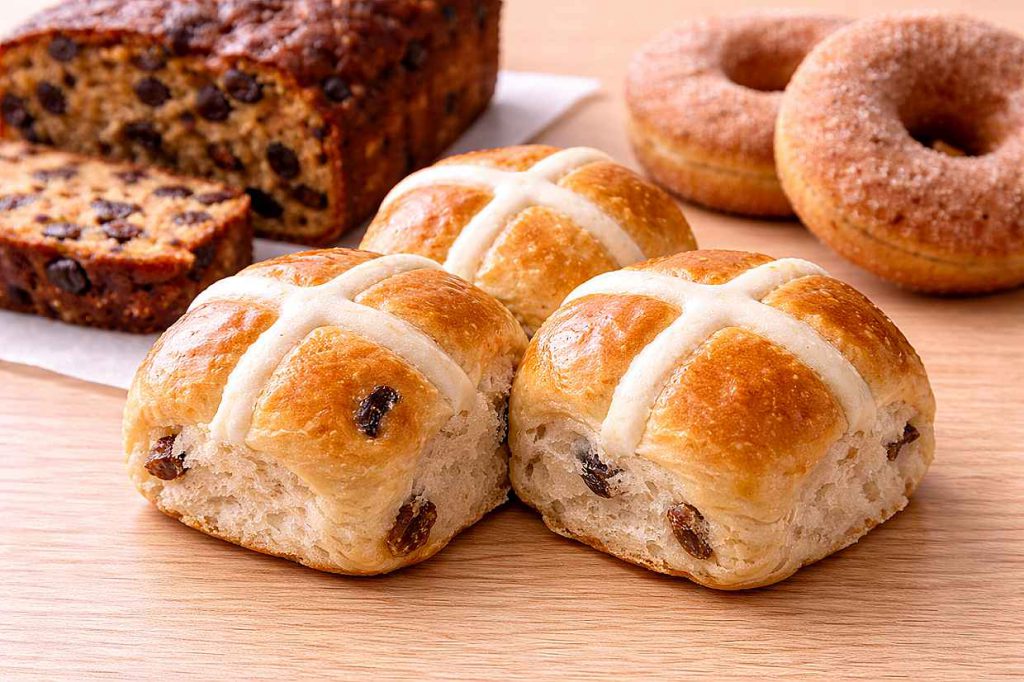 Golden hot cross buns with white crosses beside a sliced dark fruitcake and pumpkin donuts dusted with cinnamon sugar on a warm wooden surface