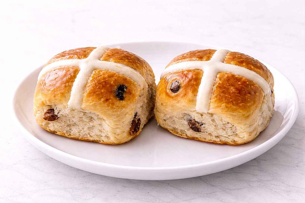 Two hot cross buns side by side on a white ceramic plate, with the left bun showing a raised baked-on flour paste cross and the right bun showing a flat white icing cross after baking