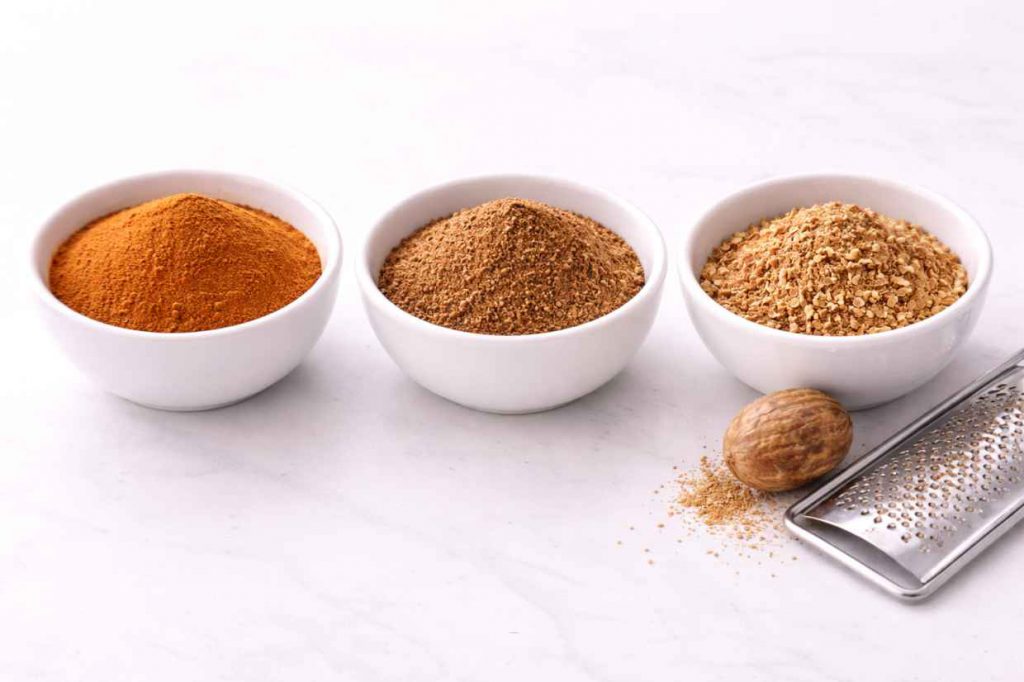 Three small white ceramic bowls containing ground cinnamon mixed spice and freshly grated nutmeg beside a whole nutmeg and grater on a white surface