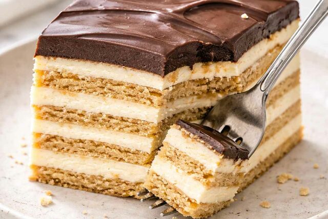 Slice of no-bake chocolate eclair cake on a speckled ceramic plate with a fork showing the distinct softened biscuit layers and creamy vanilla filling in cross-section