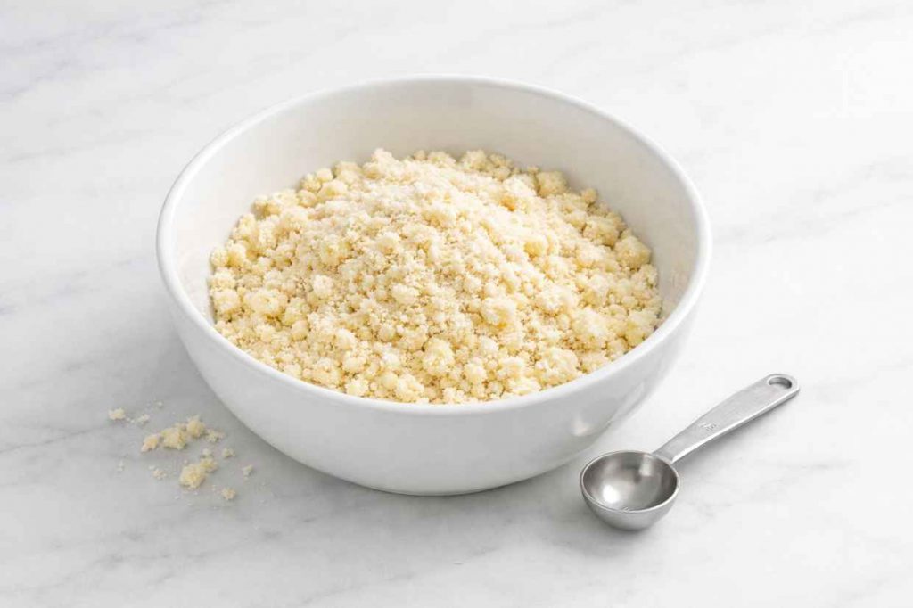 White ceramic bowl of flour and cold butter at the breadcrumb stage on a white marble surface showing the coarse sandy texture of correctly made shortcrust pastry