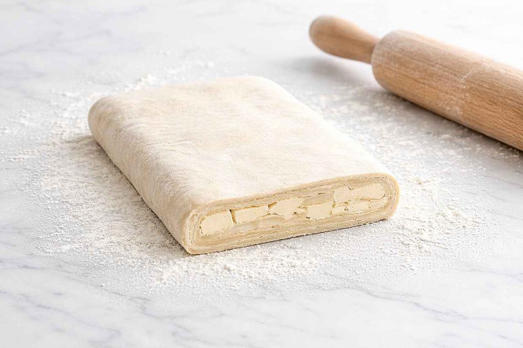Rectangle of puff pastry dough on a white marble surface with a clean cut edge showing visible cold butter pieces layered within the dough