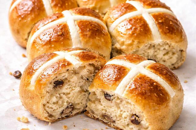 Golden hot cross buns fresh from the oven on parchment paper showing white flour paste crosses and pull-apart texture with one bun split open showing the soft spiced interior