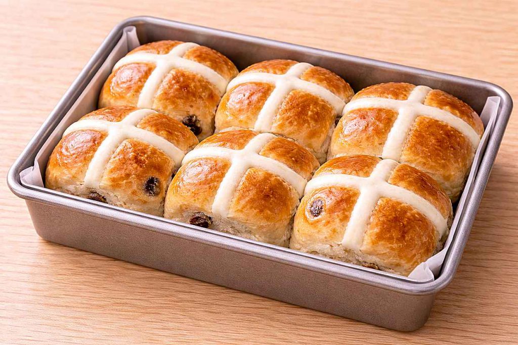 Six soft golden hot cross buns fresh from the oven with white flour paste crosses arranged in a baking tin showing pull-apart texture