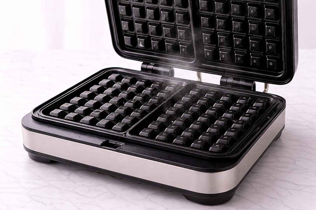 Open waffle iron on a kitchen counter with steam rising from the hot plates before adding batter showing correct preheating technique